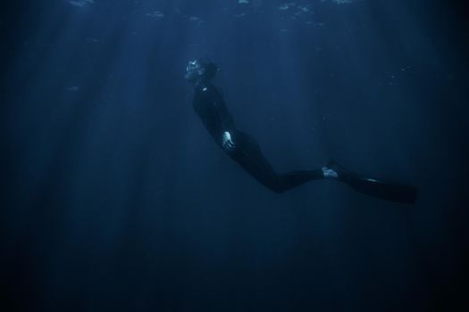 Sporty young man wearing in wetsuit and flippers snorkeling underwater in blue deep sea.