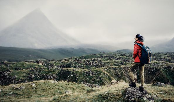 Hiker young woman with backpack walking in front of mountain in fog.