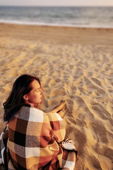 Young woman wrapped in plaid relaxing on sand beach near the sea, summer vacations.