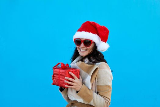 Beautiful young woman in Santa Claus hat in beige coat beret and sunglasses hold red gift over blue background