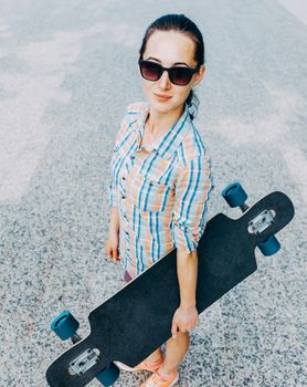 Stylish girl in sunglasses standing with longboard outdoor, looking at camera.