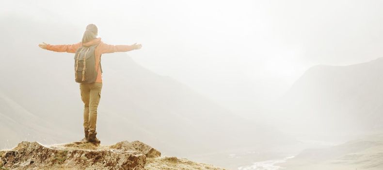 Explorer young woman with backpack standing with raised arms in the mountains, rear view.