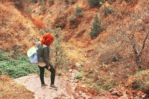 Female tourist with a backpack admires the mountain scenery. A young woman with red hair stands on the edge of a cliff. Autumn mountain landscape with rocks and trees.
