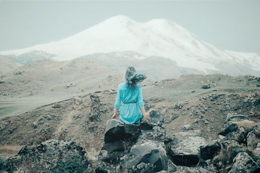 Young woman sitting on stone and enjoying view of mount Elbrus outdoor, rear view.