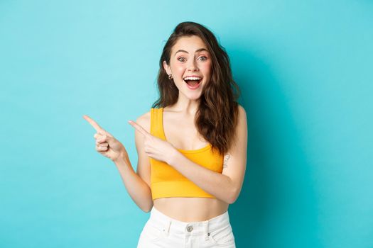 Portrait of happy young woman smiling amused, showing awesome promo deal, pointing fingers left at copy space and looking fascinated at camera, standing in summer clothes against blue background.