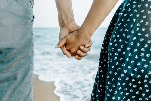 Unrecognizable couple in love holding hands together on sea coast, rear view, close-up.
