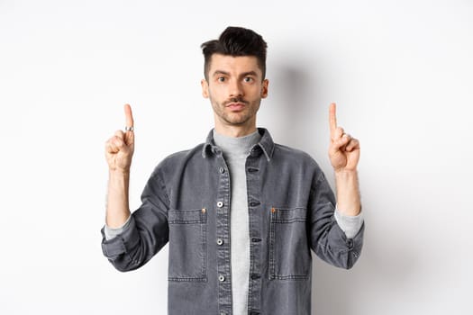 Serious caucasian guy with moustache, pointing fingers up at empty space and look at camera, white background.
