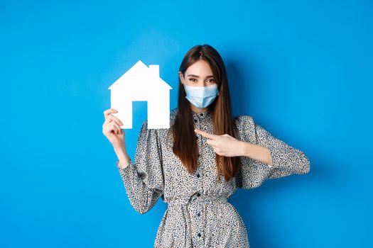 Real estate, covid-19 and pandemic concept. Smiling female model in medical mask pointing at paper house cutout, standing on blue background.
