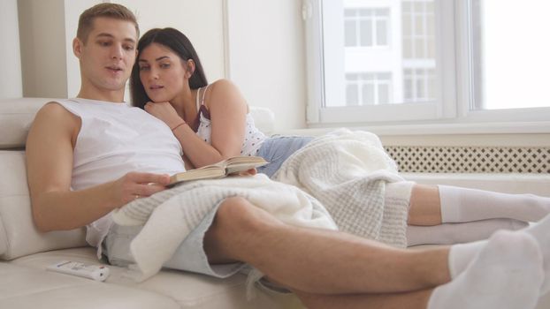 Happy young attractive couple are together on the couch, the girl reads a book, the guy lies on her lap, they're holding hands, people's attitudes