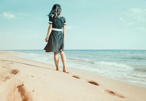 Barefoot young woman in a dress walking on sand beach leaving footprints near the sea, rear view. Summer vacations.