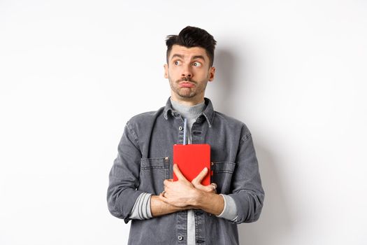 Silly young man looking aside at logo with timid face, holding his diary pressed to chest, carry journal, standing on white background.