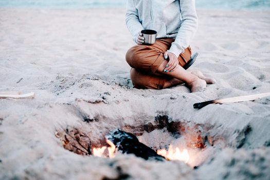 Unrecognizable woman resting near the campfire on sand beach.