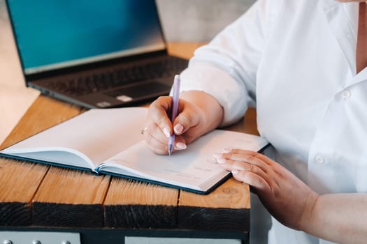 A businesswoman in a white shirt sits at a desk writing something in a notebook in the office.