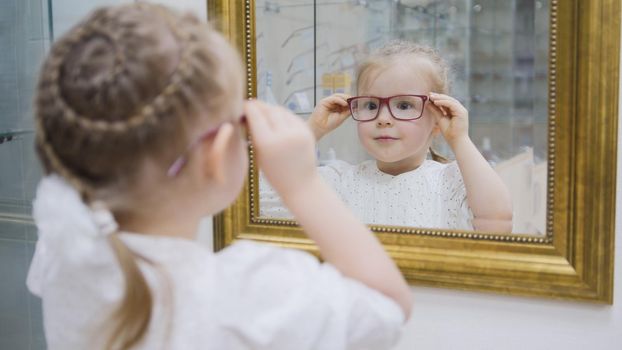 Little girl tries new glasses near mirror - shopping in ophthalmology clinic, close up