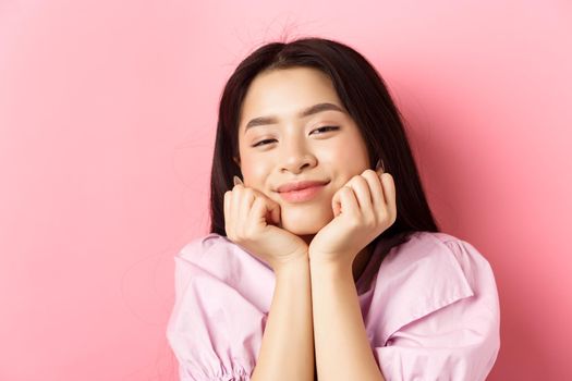 Close-up portrait of dreamy and romantic asian girl, lean face on hands and smiling, looking with admiration and happiness, standing against pink background.