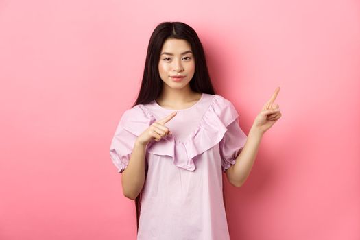 Cheeky asian girl showing interesting advertisement, pointing fingers right at logo and smiling cunning, hinting on good deal, pink background.