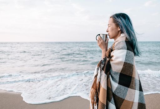 Beautiful young woman relaxing with cup of drink on sand coast near the sea, beach vacations.