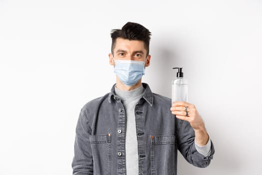 Healthy people and covid-19 concept. Young man in medical mask use preventive measures from coronavirus, showing bottle of hand sanitizer, standing against white background.