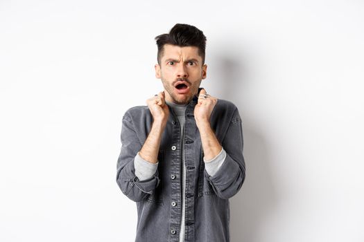 Shocked young man look confused and offended, gasping and frowning at camera, hear bad news, standing on white background.