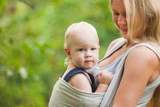 Mother is carrying her child and walking. Baby in sling outdoor.