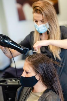 Female hairdresser drying her client's hair with a hairdryer, wearing protective masks in a beauty centre. Business and beauty concepts