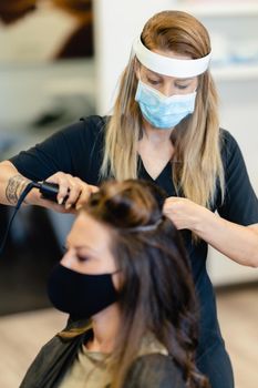 Hairdresser, protected by a mask, making waves in her client's hair with a hair iron in a salon. Business and beauty concepts