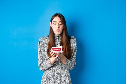 Cute birthday girl celebrating, blowing candle on cake and making wish, standing on blue background. Party and holidays concept.