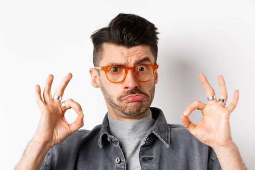 Not bad. Funny young guy in glasses pucker lips, showing okay sign and look silly at camera, standing on white background.