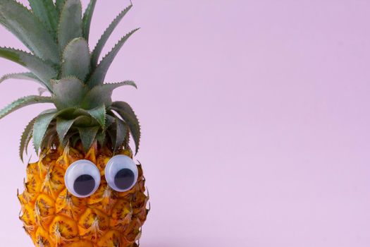 Closeup of fresh yellow funny eco pineapple with green leaves and with googly eyes on pink background, no people, enough place for text. .