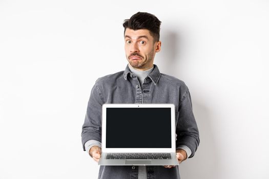 Indecisive young man pouting and shrugging while showing empty laptop screen, standing clueless on white background.