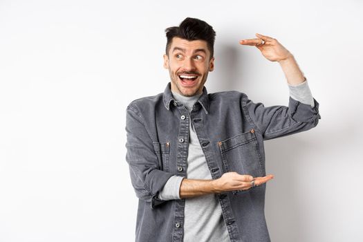 Excited guy looking at logo and showing big size thing, shaping large object and smiling amazed, standing in denim jacket on white background.