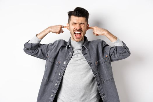 Annoyed guy shut ears and screaming, displeased by loud noise, yelling to stop noisy music, standing on white background.