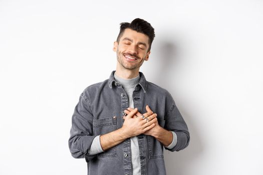 Romantic happy man holding hands on heart and smiling with closed eyes, dreaming of something, feeling nostalgic from sweet memories, standing on white background.