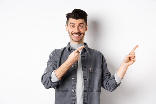 Cheerful stylish man with moustache and white smile, pointing fingers right, inviting to look here, standing on white background.