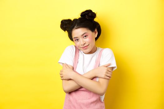 Summer lifestyle concept. Cute asian beauty girl with makeup, hugging herself and smiling romantic, standing tender on yellow background.