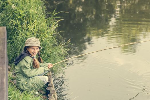 little fisher girl sitting on a river bank with a rod smiling