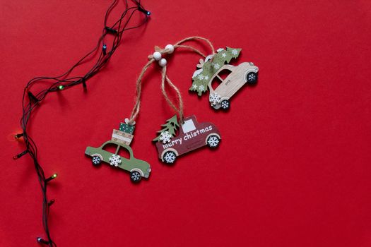 Three wooden car toys for Christmas tree near colocful light bulb on red background, top view, copy space.