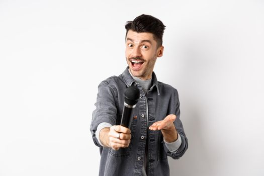 Handsome smiling man stretch out hand with microphone, giving you mic, standing on white background.