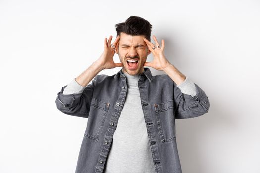 Frustrated young man shouting and touching head distressed, standing bothered by loud noise or headache, standing on white background.