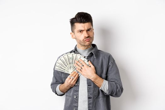 Greedy guy frowning and hugging dollar bills, unwilling to share money, standing on white background.