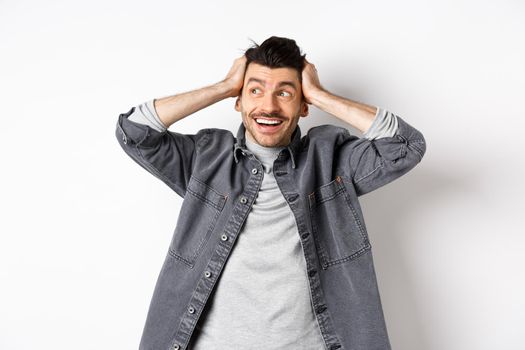 Excited young man looking relieved and happy aside, smiling and staring at logo, holding hands on head cheerful, standing on white background.