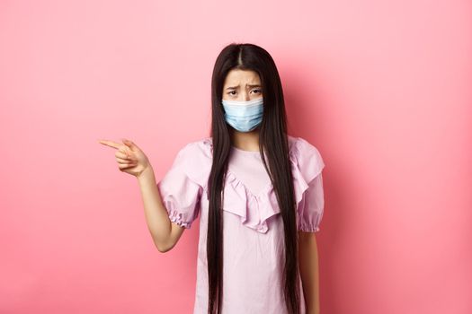 Covid-19, pandemic lifestyle concept. Sad and gloomy asian girl in medical mask pointing left, complain on unfair situation, standing disappointed against pink background.