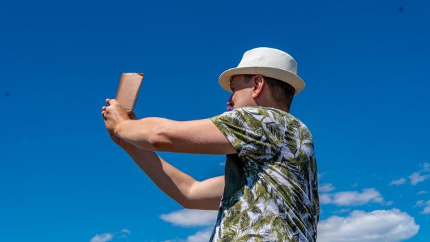The guy takes pictures on the blue background of the sky and white clouds, in the afternoon of summer in a white cap on the phone