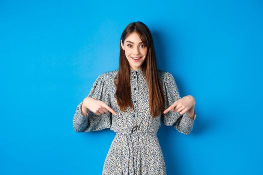 Excited caucasian woman in dress pointing fingers down at logo, showing fantastic news and smiling, standing on blue background.