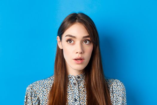 Close-up portrait of surprised young woman with natural beauty, looking at upper left corner and gasping, standing on blue background.