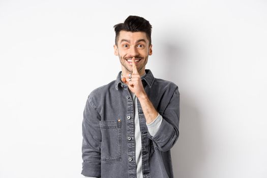 Happy guy shushing at camera, smiling with finger on lips, asking to keep quiet, telling big secret or making surprise, standing on white background.