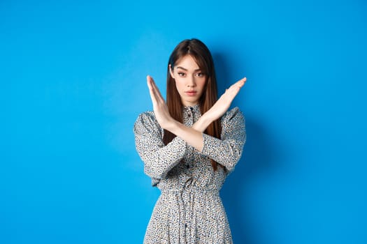 Serious caucasian woman in dress tell no, show cross stop gesture and looking confident, making warning sign, prohibit something bad, standing on blue background.