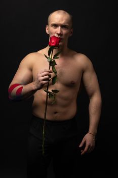 Athletic man holding a red rose, naked for the day of velentin pumped up, on his arm a teip of a bodybuilder romantic. lover guy LGBT. In the studio on a black background with a wonderful smile