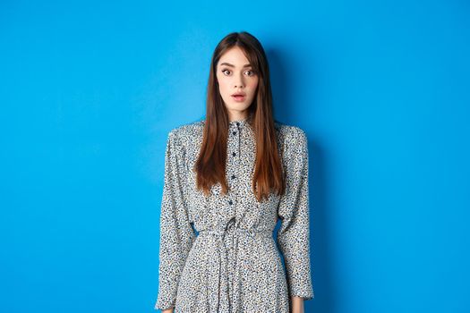 Shocked caucasian woman with long hair, wearing dress, gasping and standing in stupid against blue background, stare with disbelief.