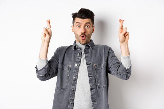 Excited young man cross fingers for good luck and gasping, holding breath as waiting important news, begging or praying, standing on white background.